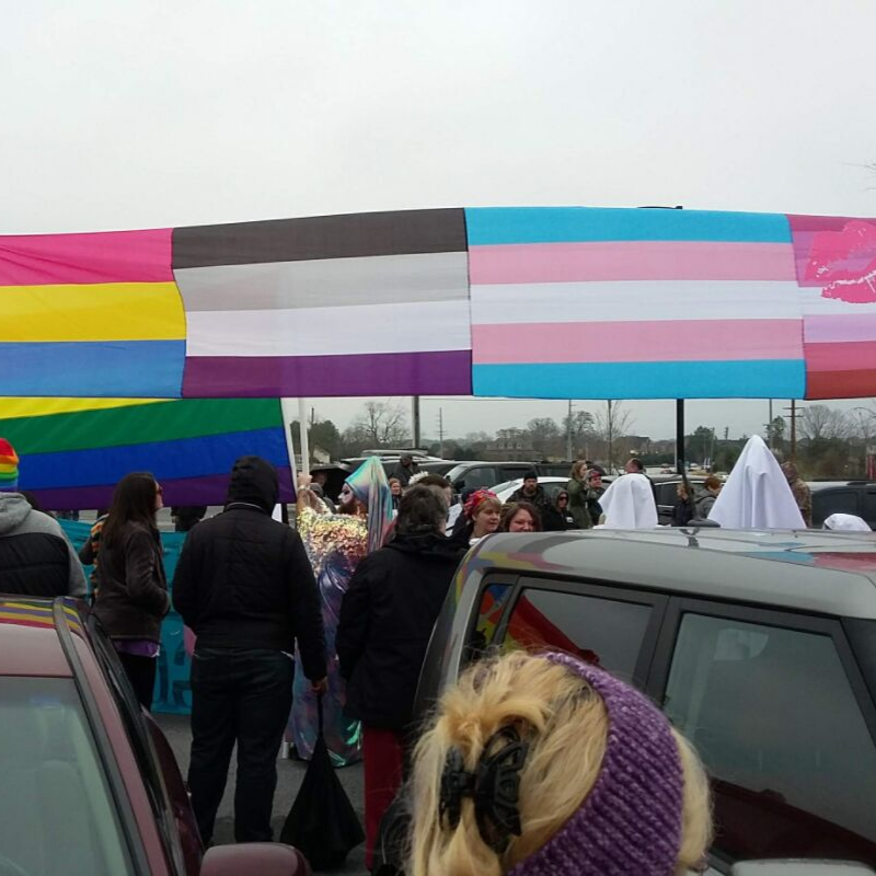 A banner of LGBTQIA+, pansexual, transgender, asexual, & lesbian Pride flags hangs over the Drag Queen Story Hour protest.